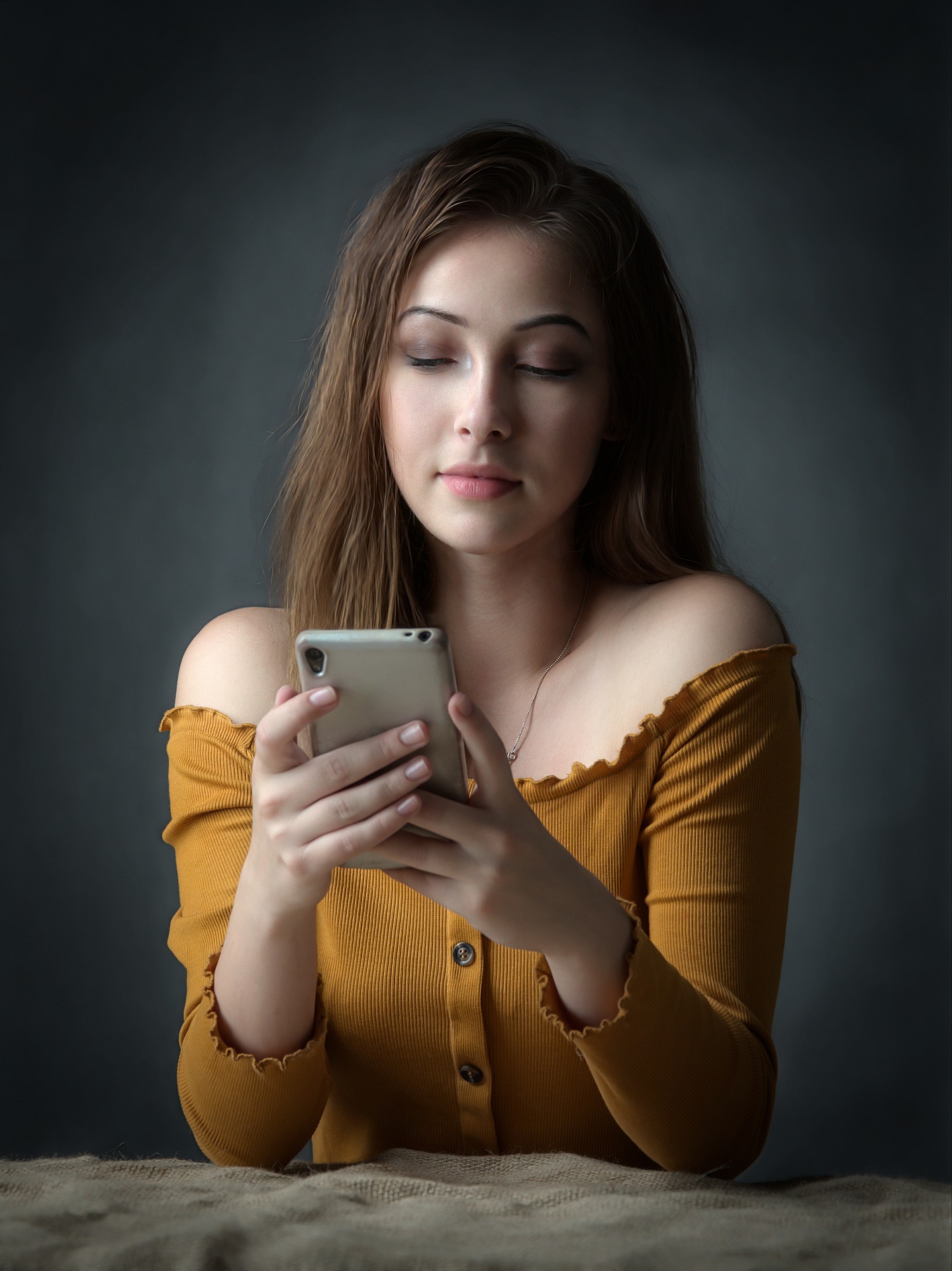 Ein junges Mädchen mit dunklem Hintergrund sitzt in einem senffarbenen T-Shirt mit nackten Schultern hinter einem Tisch und schaut auf ein Mobiltelefon. Bild zum Thema: Filter als Schönheitsideal: Snapchat Dysmorphia