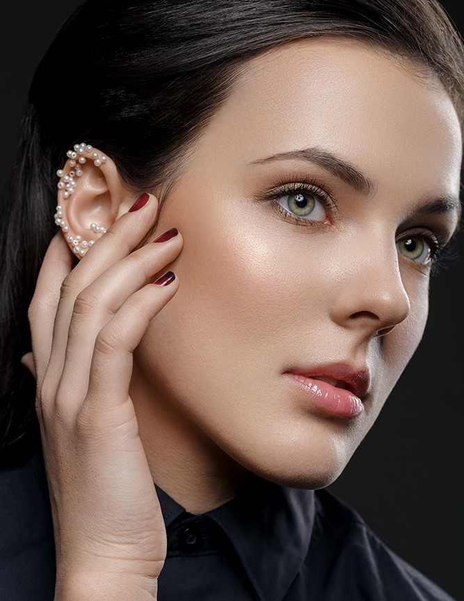 Schöne junge Frau mit natürlichem Make-up und aufgeklebten Perlen am Ohr trägt ein schwarzes Hemd. Schönheitsaufnahme auf schwarzem Hintergrund. Bild zum Thema Ohrenkorrektur